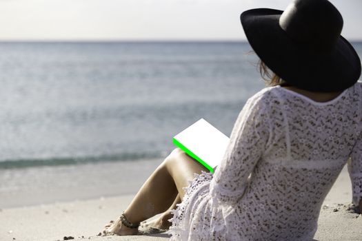 Young woman dressed in a white lace dress, white underwear and big black hat from behind sitting by the sea holding a book with blank cover on her legs