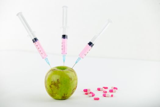 Concept: human GMO manipulation of nature and relative poisoned fruits. Close-up of an apple contaminated with three syringe threaded in and medicines on a white background