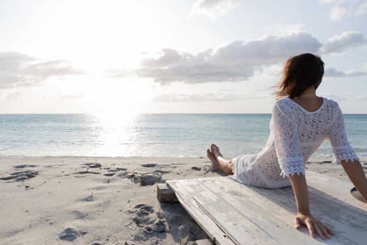 Young woman from behind sitting by the sea looks at the horizon at dawn in the wind, dressed in a white lace dress and white underwear and long hair