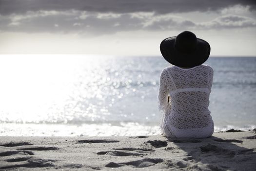 Young woman with long hair from behind sitting by the sea looks at the horizon at dawn in the wind, dressed in a white lace dress, white underwear and large black hat