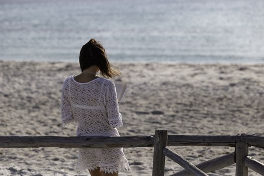 Young woman from behind reads a book on the beach looking at the horizon at dawn in the wind, dressed in a white lace dress, white underwear and long hair