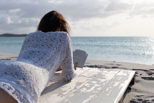 The young woman from behind lying by the sea looks at the horizon at dawn in the wind, dressed in a white lace dress and white underwear and long hair