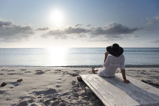 Young woman with long hair from behind sitting by the sea looks at the horizon at dawn in the wind, dressed in a white lace dress, white underwear and large black hat