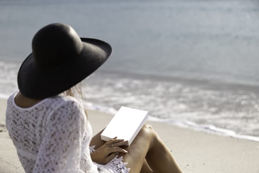 Young woman dressed in a white lace dress, white underwear and big black hat from behind sitting by the sea holding a book with blank cover on her legs