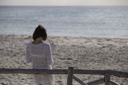 Young woman from behind reads a book on the beach looking at the horizon at dawn in the wind, dressed in a white lace dress, white underwear and long hair