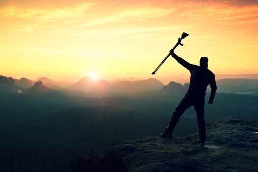 Tourist with forearm crutch above head achieved mountain peak. Hiker with broken leg in immobilizer and medicine  poles hold hand in air. Colorful misty valley bellow silhouette.