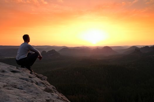 Young hiker in black trousers and grey shirt  sit on cliff's edge. Tourist watching sunrise above  misty hilly valley bellow mountain