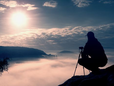 Amateur photographer takes photos with mirror camera on peak of rock. Dreamy fogy landscape, spring orange pink misty sunrise in a beautiful valley below