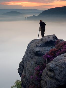 Professional photographer takes photos with mirror camera and tripod on peak of rock. Dreamy fogy landscape, spring orange pink misty sunrise in beautiful valley below.