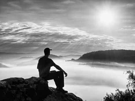 Alone hiker in black trousers and red shirt  and red cap sit on cliff's edge and looking to dreamy mist bellow. Hilly landscape with marvelous valley 