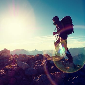Lens flare light, strong defect. Silhouette of man with hood, backpack and poles in hand. Man walk on rocky peak of mountain. Sunny day rocky mountains.