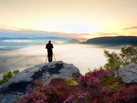 Professional photographer takes photos with mirror camera and tripod on peak of rock. Dreamy fogy landscape, spring orange pink misty sunrise in beautiful valley below.