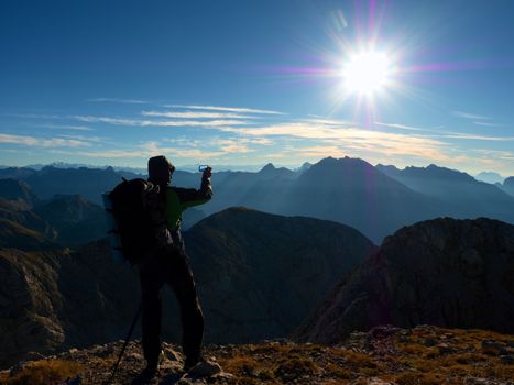 Experienced hiker takes phone photo. Man on Alps mountain peak.View to blue sky above deep foggy valley. Mountains increased from humidity