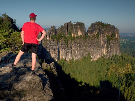 Adult hiker in red shirt and dark pants. Tall man on the peak of sandstone cliff watching down to landscape.