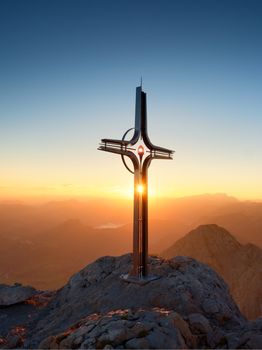 Steel crucifix at mountain peak in Alps. Sharp rocky summit, daybreak Sun in sky. Cross raised in memory of victims of mountains. Vivid photo.