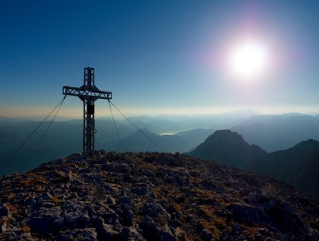 Steel crucifix at mountain peak in Alps. Sharp rocky summit, daybreak Sun in sky. Cross raised in memory of victims of mountains. Vivid photo.