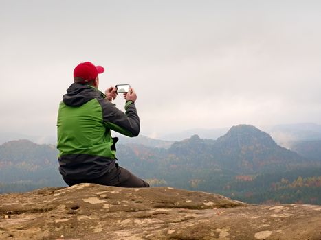 Man takes photos with smart phone on peak of rock empire. Melancholy fogy landscape, spring orange pink misty sunrise in a beautiful valley of rocky mountains.