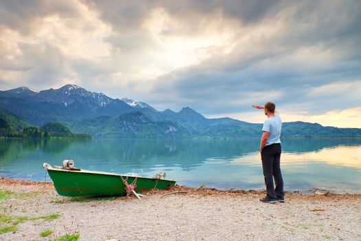 Adult man in blue shirt walk at old fishing paddle boat at mountains lake coast. Vintage photo effect
