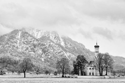 Old church with high bell tower and graveyard bellow mountains.  April weather. Wet spring snow in already fresh green grass  of meadow bellow mountains. 