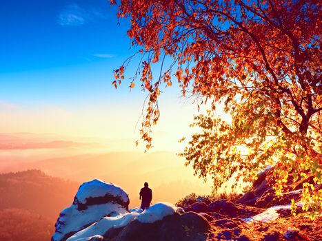 Sunny morning with first snow. Photographer preparing camera on tripod. Snowy rocks, in valley bellow colorful leaves forest. View over misty and foggy valley to Sun.