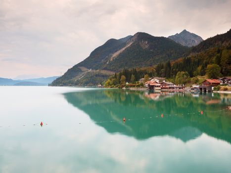 Village on the bank of Alps lake. High mountain peaks in mirror of gren water level. Beginning of autumn mountains.