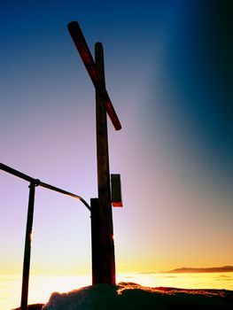 Modest wooden cross raised  on rocky mountain summit . Sharp snowy peak. Daybreak Sun in sky. Wooden unpretentious crucifix in memory of victims of mountains. Vivid photo.