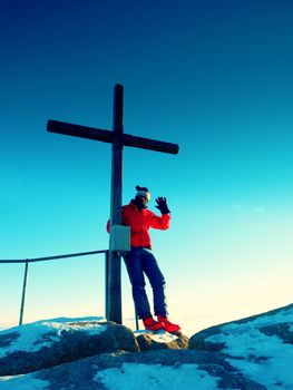 Tourist in red blue winter clothes on sharp summit  with modest cross.  Winter Sun in sky. Wooden unpretentious crucifix  raised  on mountain peak in memory of victims of mountains.