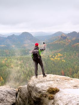 Tourist with backpack stayon cliff and  takes photos with smart phone of rainy valley. Melancholy atmosphere in foggy valley below