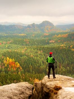 Tall adult  photographer prepare camera for taking picture of fall  mountains. Photograph at daybreak above  colorful  valley. Landscape view of autumn mountain hills and hiker above