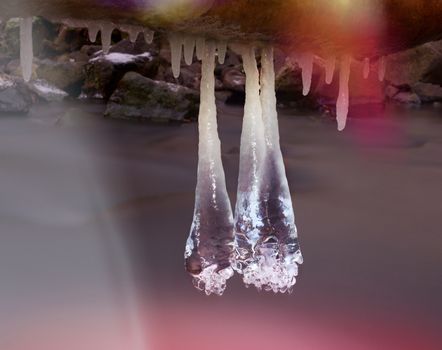 Lens defect. Long icicles hang above dark freeze  water of mountain stream. Winter season at river, thin icicles are hanging on fallen trunk above milky water level. 