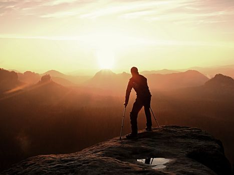 Bended man with broken leg and medicine crutch.  Hiker with leg in immobilizer achieve peak of mountain. Deep misty valley bellow silhouette.  Spring daybreak