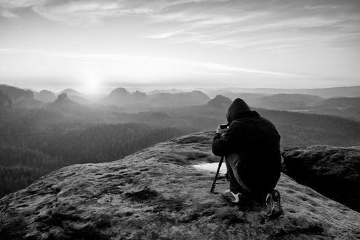 Professional on cliff. Nature photographer takes photos with mirror camera on peak of rock. Dreamy fogy landscape, spring orange pink misty sunrise in a beautiful valley below. 