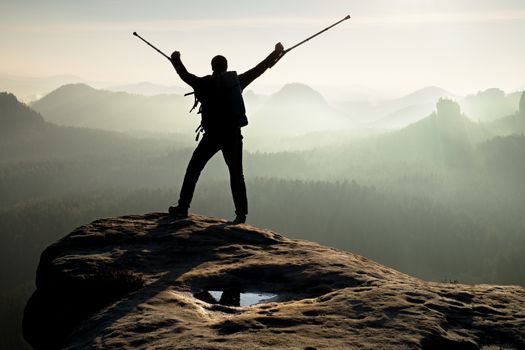 Hiker with broken leg in immobilizer. Tourist with  medicine crutch above head achieved mountain peak. Deep misty valley bellow silhouette of man with hand in air. Spring daybreak