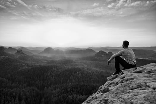 Young hiker in black trousers and grey shirt  sit on cliff's edge. Tourist watching sunrise above  misty hilly valley bellow mountain
