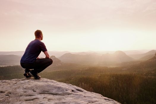 Tourist in squatting position on peak of sandstone rock and watching into colorful mist and fog in morning valley. Orange daybreak at horizon