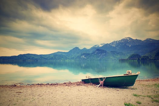 Abandoned fishing paddle boat on bank of Alps lake. Morning lake glowing by sunlight. Dramatic and picturesque scene. Mountains in water mirror.