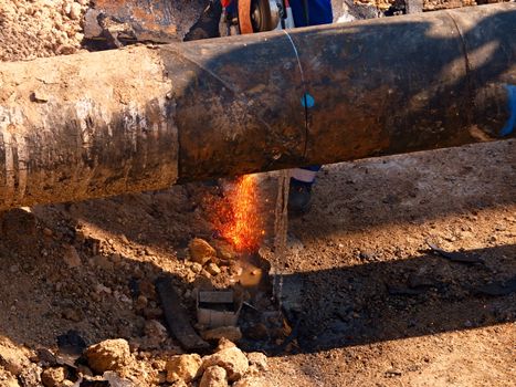 Worker staff is cutting big metal tube with hand grinder. Burning parks are flying down to wet clay. Staff use safety clothes, protective boots. Deep excavation pit.