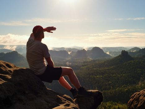 Man sit on the peak of rock. Hiker shadowing eyes with raised arm,  watching into colorful mist and fog in forest valley.