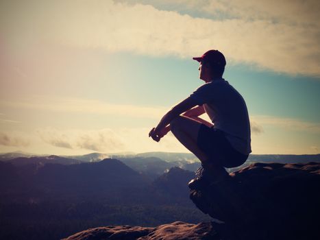 Hiker man take a rest on mountain peak. Man sit on summit, bellow spring valley. Bright morning Sun shining in sky. 