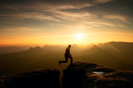 Hiker in black is jumping between mountains peaks. Wonderful daybreak in rocky mountains, heavy orange mist in deep valley. 