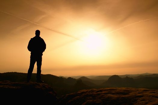 Sportsman in black with hands in pockets stand on the peak in rock empires park and watching over the misty and foggy morning valley to Sun. Silhouette of man.