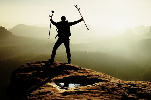 Hiker with broken leg in immobilizer and medicine pole above head achieved rocky summit. Deep misty valley bellow sharp edge. Silhouette of tourist with hand in air. Spring daybreak