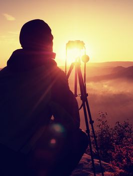 Amateur photographer takes photos with mirror camera on peak of rock. Dreamy fogy landscape, spring orange pink misty sunrise in a beautiful valley below