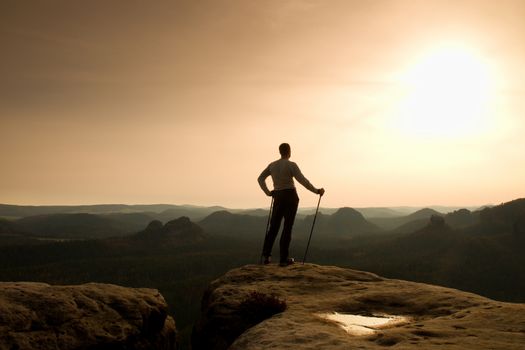 Ginger hair man in grey t-shirt and dark trekking trousers on sharp rock. Tourist with pole above misty valley. Sunny misty moment in rocky mountains. 