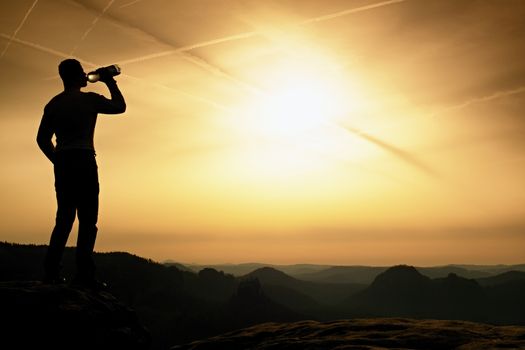 Thirsty hiker in black pants with bottle of water. Sweaty tired tourist on the peak of sandstone rocky park Saxony Switzerland watching into misty landscape.