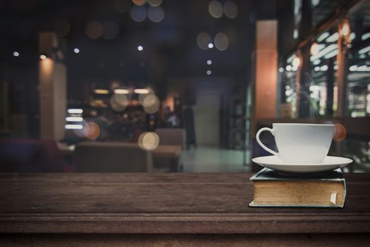 Hot cup of coffee on big book on wood table in front of blurred cafe restaurant background, copy space can be placed your product.