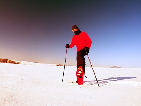 Man cleaning snowshoe. Winter tourist with snowshoes walk in snowy drift. Sunny freeze weather. Hiker in pink sports jacket and black trekking trousers snowshoeing in powder snow.