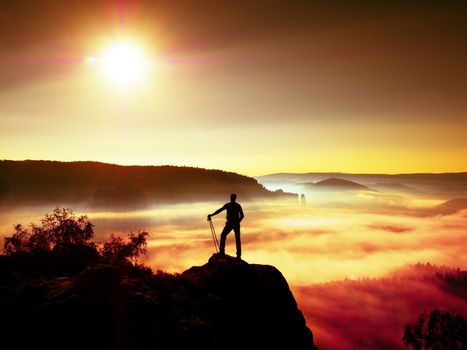 Tall backpacker with poles in hand. Sunny misty daybreak in rocky mountains. Hiker with backpack stand on rocky view point above valley. Vivid and strong vignetting effect.