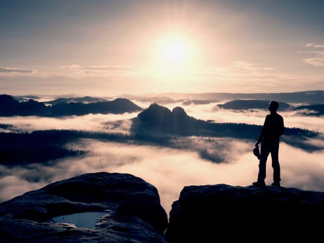 Hiker silhouette stand on cliff and watching over misty valley to Sun. Blue filtered photo. Tourist on edge. 