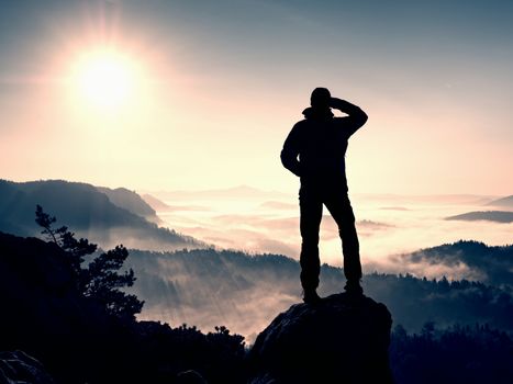 The man standing high on cliff. Hiker climbed up to rocky peak and enjoy view to valley bellow. Man  watch over misty and foggy morning valley in morning Sun. 